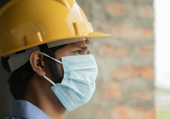 Head shot side view of Construction worker seeing outside from construction sites or industry window - construction worker in a construction helmet with medical mask due to coronavirus or covid-19 crisis