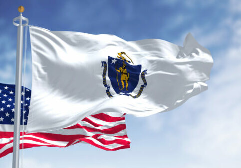The Massachusetts state flag waving along with the national flag of the United States of America. In the background there is a clear sky. Massachusetts is a state in the New England region of the United States